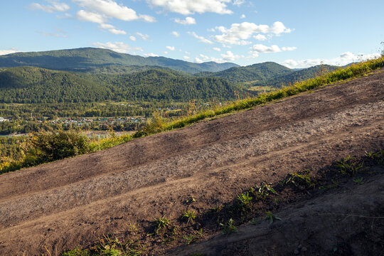 Katun river valley summer landscape photo with a sunlit dirt road, offering a panoramic view of a valley with a small village and forested mountains under a blue sky