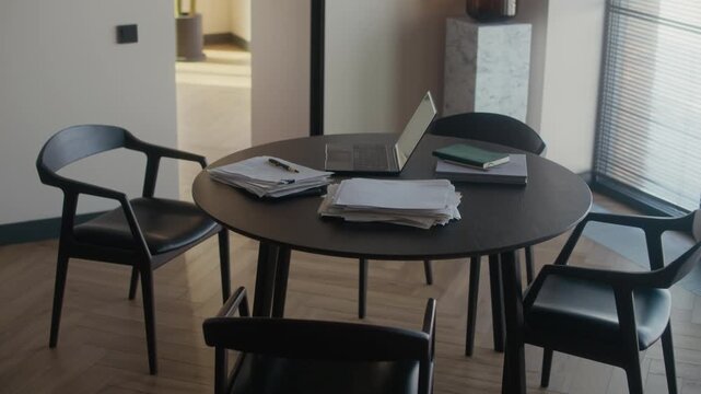 Laptop and pile of printed novel drafts on dark wooden round table with chairs in living room interior, writer workspace at home