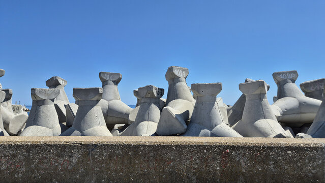 A row of concrete tetrapods, used as breakwaters, sit behind a low wall under a clear blue sky. Each numbered block features four protruding legs designed to interlock and dissipate wave energy.