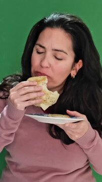 Middle-aged woman with dark hair savoring a delicious flatbread sandwich against a green screen background, taking large, enjoyable bites