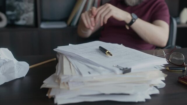 Unrecognizable male author sitting at desk with crumpled paper and pile of novel drafts in foreground, warming up and stretching his hands