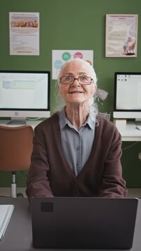 Vertical shot of elegant Caucasian senior woman using computer in retirement home and looking up at camera with smile, story format