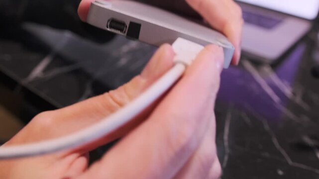 Close-up of hand connecting cable to external SSD drive, user plugging data cable into storage device port for file transfer and backup on workspace desk. Connecting external SSD cable to device