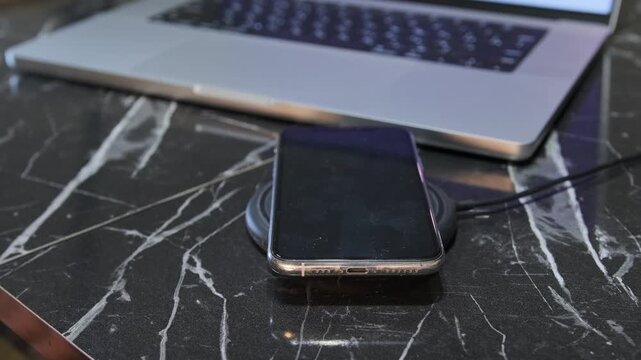 Hand placing smartphone on wireless charging pad, close-up of user setting device for contactless charging near laptop workspace on desk. Wireless charging phone