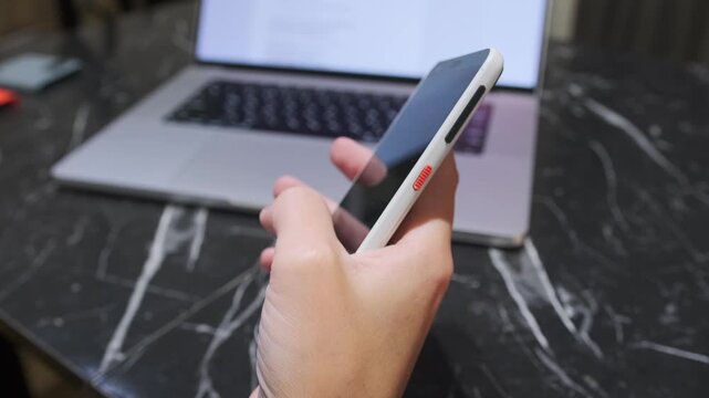 Hand pressing smartphone power button to turn screen off, close-up of locking mobile device during everyday usage near laptop on desk. Pressing screen off button on phone