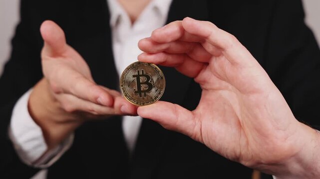 Une main tient une pi&egrave;ce de Bitcoin dor&eacute;e entre deux doigts. En arri&egrave;re plan, une personne en costume et chemise blanche.
