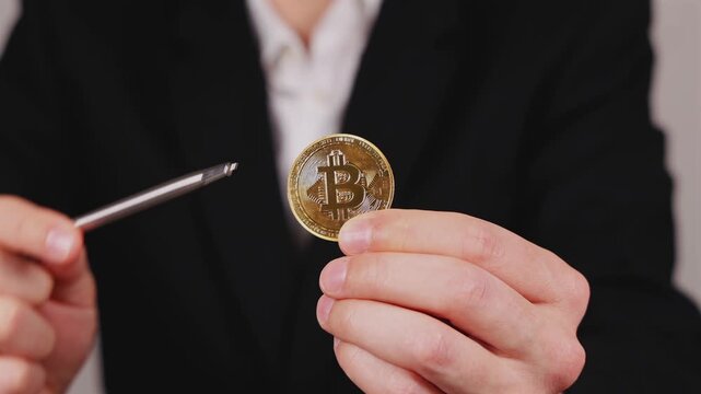 Une main montre une pi&egrave;ce de Bitcoin dor&eacute;e avec un stylo. En arri&egrave;re plan, une personne en costume et chemise blanche. 