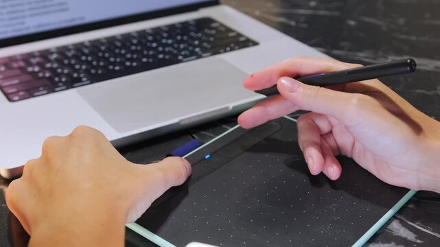 Hand drawing and pressing shortcut buttons on graphic tablet with stylus pen, close-up of digital artist working on illustration and design near laptop workspace. Drawing on graphic tablet with stylus