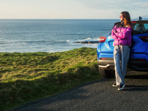 Young woman in colorful jacket is standing by a blue rented car and looking at stunning nature scenery with rough ocean coast at sunset. Mullaghmore, county Sligo. Travel and tourism in Ireland.