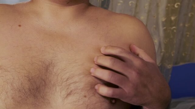 man scratching chest skin close-up view, hand rubbing irritated area on torso, expressing itching and discomfort sensation, dermatology symptom and body care concept in indoor home setting