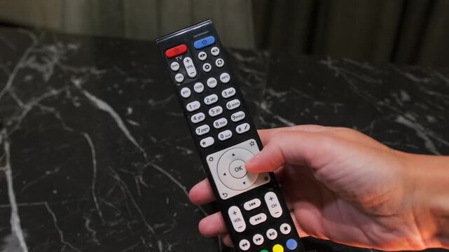Hand pressing button on TV remote control, close-up of user clicking channel or volume controls while operating home entertainment device. Using TV remote control