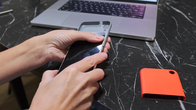 Hands putting protective armored case on smartphone, close-up of device protection setup, user installing durable cover to prevent damage and improve grip on desk near laptop