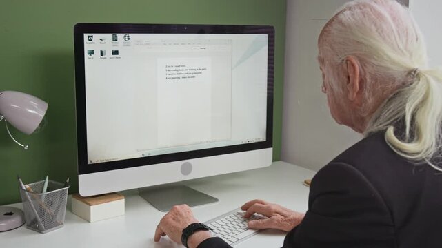 Over shoulder shot of white haired senior man using PC in library or computer class and slowly typing on keyboard entering text into digital document, copy space