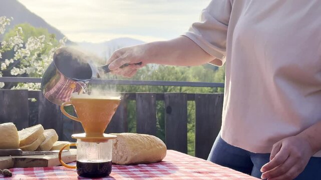 Pouring hot water over coffee using pour over brewing method on rustic outdoor terrace with fresh bread and scenic mountain background. Manual coffee preparation in natural sunlight and slow living 