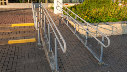 Inclined concrete accessibility ramp featuring gray metal safety handrails stands beside stone stairs with yellow warning stripes providing barrier free entry for disabled people © Dmitry