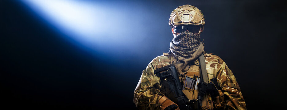 Soldier in full gear holding assault rifle on dark smoke background. Concept defense and resilience.
