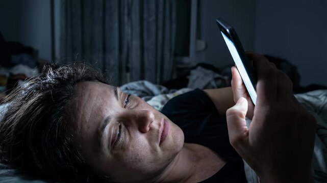 Tired woman lying in bed scrolling on her smartphone late at night, her face illuminated by the bright screen light which disturbs her sleep and causes digital eye strain