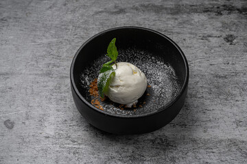 Scoop of vanilla ice cream served in black ceramic bowl with mint leaf garnish, powdered sugar and crunchy flakes on gray stone background © sutulastock