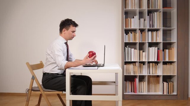 Man in a striped shirt and tie sitting at a white desk, typing on a laptop and holding a red apple, promoting healthy remote work and productivity in a home office setting