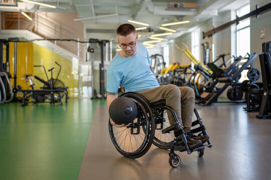Disabled man in wheelchair working on upper body and arms strength with medicine ball on rehabilitation gym training. Male sportsman doing power exercises using heavy slam ball to built strong muscles