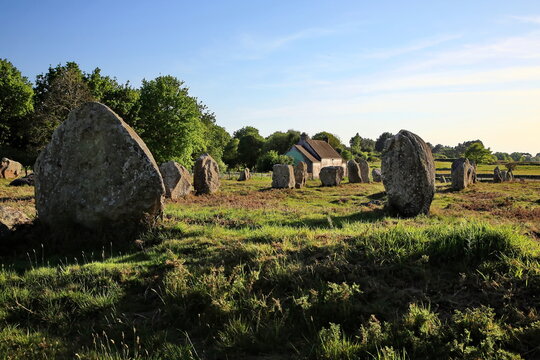 Antike Menhire erstrecken sich in langen Reihen &uuml;ber eine gr&uuml;ne Wiese vor einem einsamen Steinhaus unter strahlend blauem Himmel im warmen Abendlicht der Bretagne.