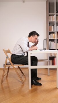 Vertical video man concentrating on laptop at desk with slumped posture, highlighting ergonomic risk and potential back pain in workspace