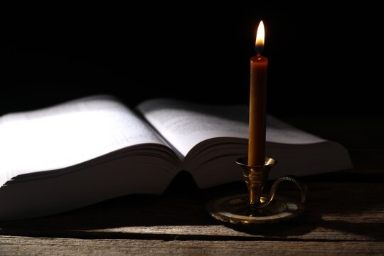 Burning candle and Holy Bible on wooden table in darkness, closeup