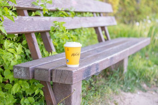 yellow McCaf&eacute; coffee cup with logo on wooden bench in park, blurred natural backdrop, modern mobility, outdoor lifestyle, fast food beverage, branded cup, ideal ads about travel, breaks consumerism