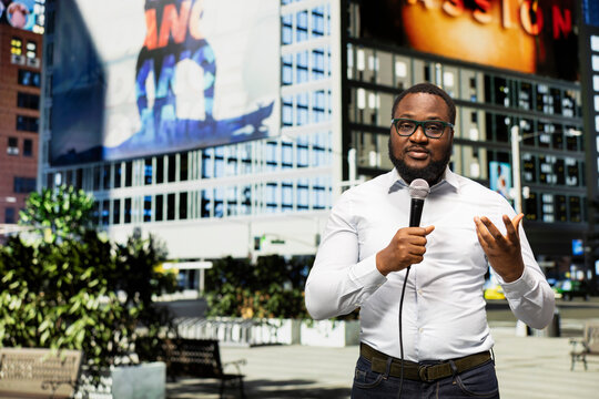 Black field journalist reports live with microphone on downtown street, conveying important information and press commentary. Maintaining professional presence in busy urban background.
