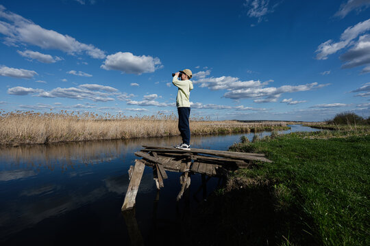 Young person observing marshland with binoculars from a rickety wooden dock under a vast blue sky with scattered clouds