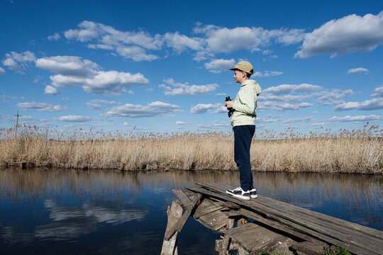 Boy on a weathered wooden dock holding binoculars overlooking a marsh pond under a blue sky with clouds, nature exploration and outdoor adventure