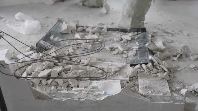 Empty room during renovation. Construction debris, dust, and rubble cover concrete floor. Bags of waste stand near unpainted walls. Natural light from windows illuminates messy, unfinished interior