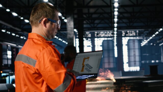 Engineer operates automation software on a laptop to control robot equipment in fabrication and construction tasks. Industrial power and precise engineering development in industry plant.