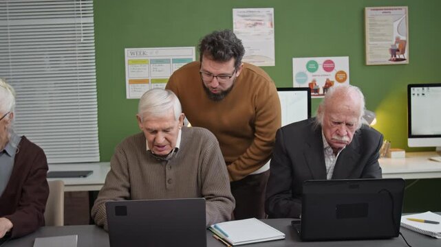 Following front view shot of caring male teacher overseeing group of seniors using laptops during computer literacy class and assisting adult students one by one