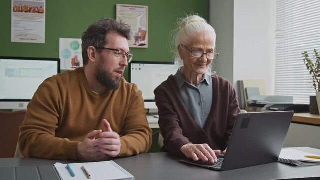 Medium shot of smiling male teacher assisting white haired senior woman using laptop in computer class or library and giving advice providing personal mentorship, copy space