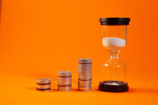 Time is money concept. Hourglass and coins on orange background.