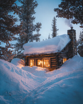 Traditional wooden log cabin in a remote winter wilderness. The cabin is illuminated by golden interior light, surrounded by high snowdrifts and evergreen trees under a twilight sky.