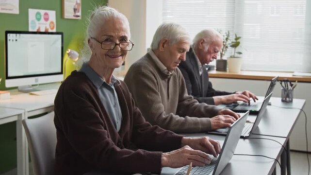 Side view shot of white haired senior woman using laptop in computer literacy class and turning looking at camera with smile while sitting in row at desk with group of adult students, copy space