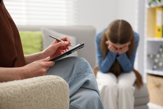 Professional child psychologist working with upset girl and writing in clipboard on sofa indoors, closeup