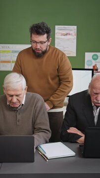 Vertical shot of supportive male teacher assisting senior people using laptops in retirement home, social media story format