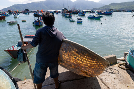 A fisherman carries a coracle boat and oar at a busy harbor in Hon Mieu, Nha Trang, Vietnam. Many fishing boats are moored in the bay.