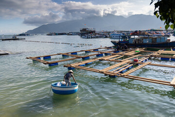 A fisherman in a traditional conical hat works on a floating fish farm in the waters of Hon Mieu, Nha Trang, Vietnam. The man is in a round boat, tending to the wooden structures. © Zenstratus