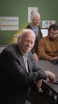 Vertical shot of white haired senior man looking at camera smiling while sitting at desk in school classroom, elderly outreach program