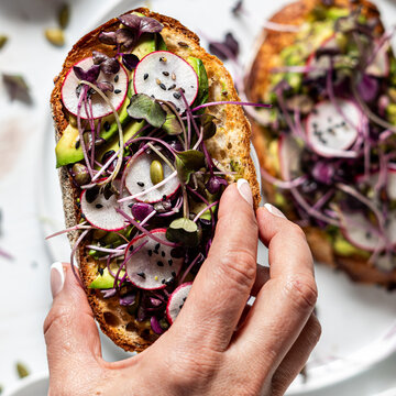 A hand holding a slice of avocado toast with raw ingredients and another in soft focus below.