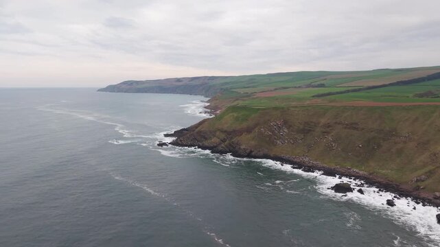 Cinematic aerial sweep of rugged vertical cliffs and crashing waves, Scottish coastline.