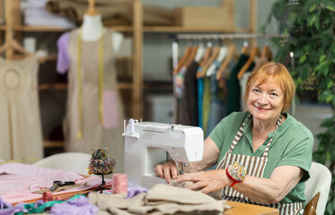 Fototapeta premium Mature female seamstress is sitting near sewing machine and create new dress in the workshop