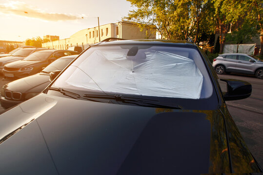Metallic foil sun protector. Sedan parked on city street during sunny day, windshield sunshade. Dark car in parking area with bright sun flare, use of reflective windshield cover for heat protection.
