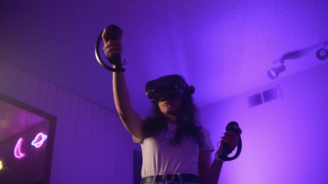 Young woman wearing VR headset raising controllers interacting with virtual reality entering digital space and controlling gameplay in modern technological environment