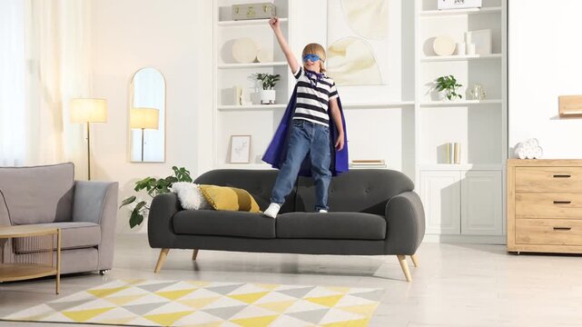 Little boy in superhero costume jumping on sofa and pretending to fly at home