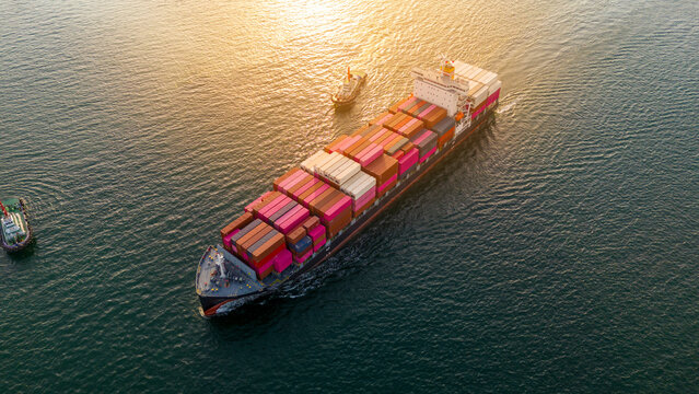 cargo container ship sailing on the sea at sunset. Two tugboats are guiding the heavy vessel through the golden water. Global maritime trade, freight transport and logistics.